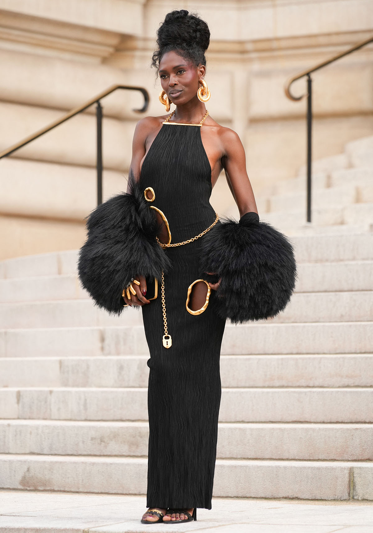 Jodie Turner-Smith looks like a goddess in her black viscose knit dress with a tree-bark texture, irregular cutouts with hammered gold trims, and chain accents around the waist and neck