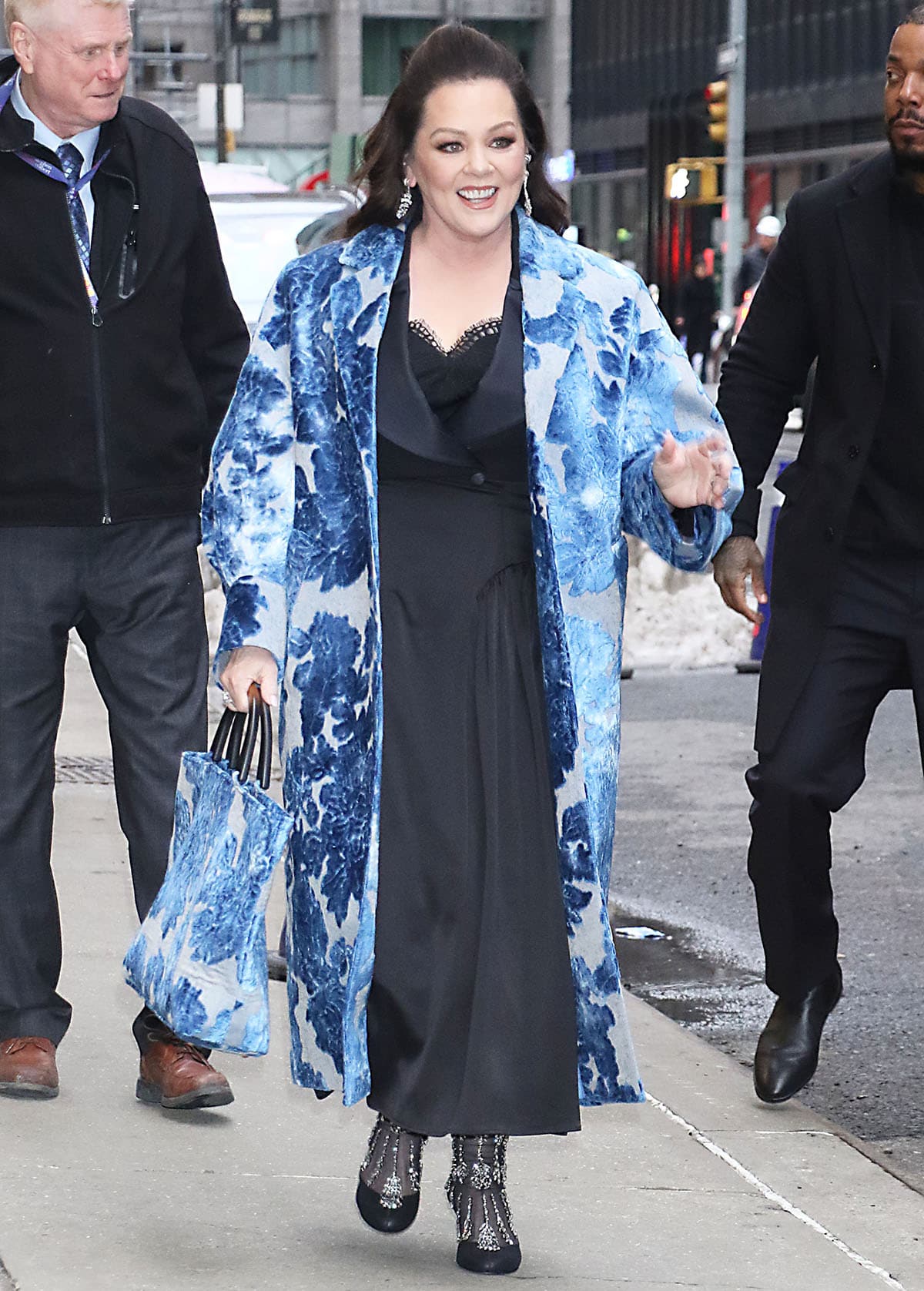 Melissa McCarthy pairs her already ornate crystal mesh boots with a striking blue floral jacquard coat, a matching tote bag, and a black satin blazer dress underneath