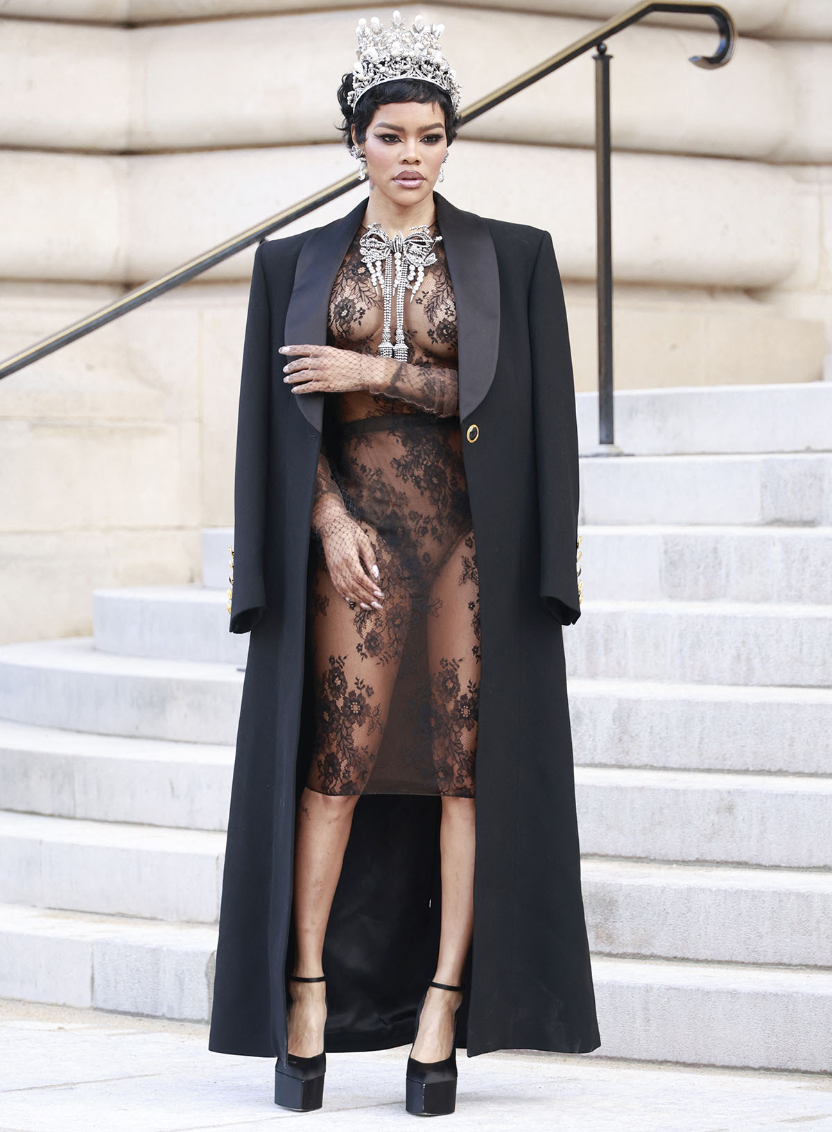Teyana Taylor pairs her simple platform pumps with a commanding see-through sheer lace top and mini skirt, a large silver chandelier brooch, a long coat, and a crown