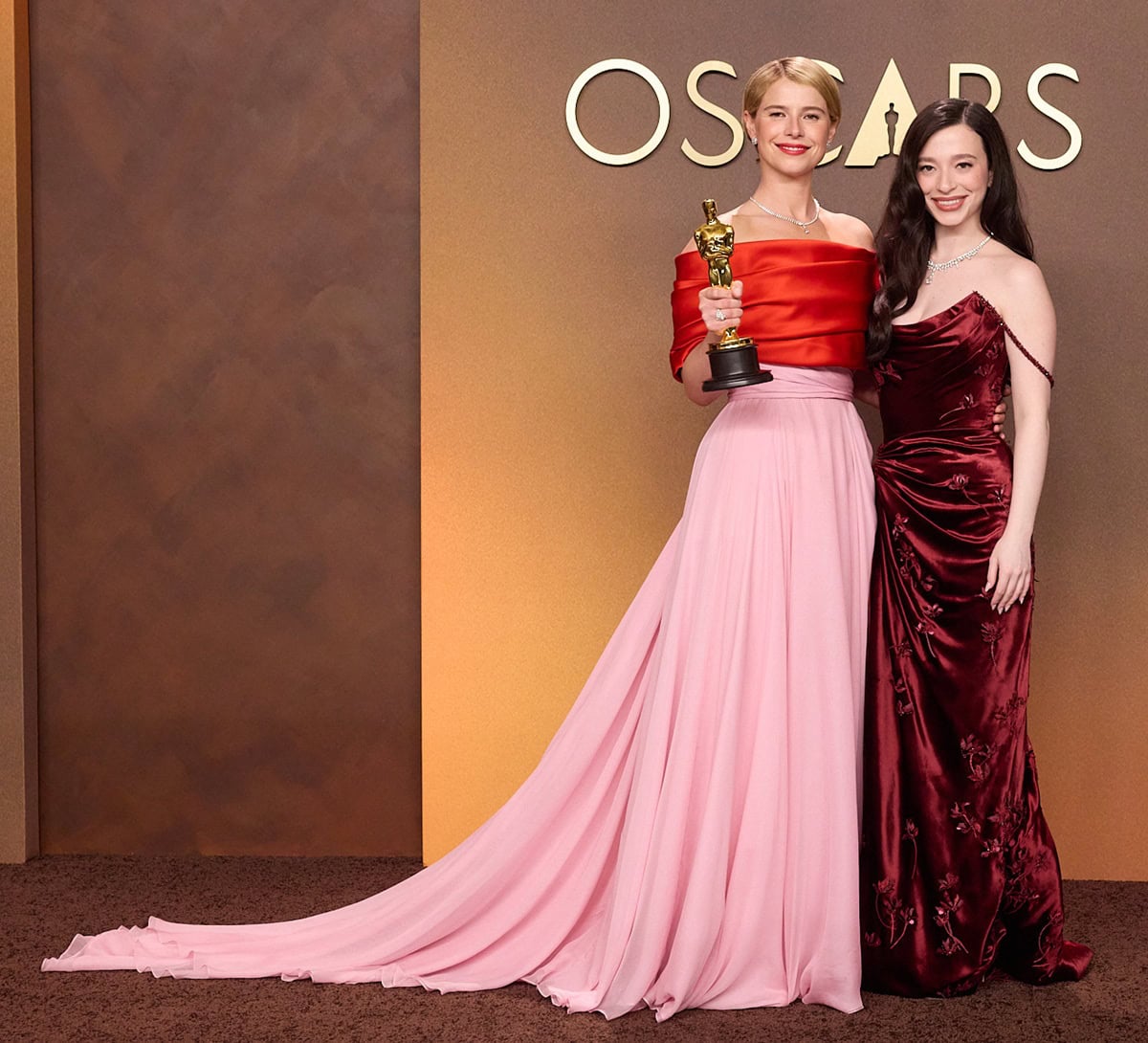 Jessie Buckley poses with her Oscar for Actress in a Leading Role beside Mikey Madison, who stuns in a rich red velvet Dior gown with floral appliqués, a thigh-high slit, and delicate beaded off-the-shoulder straps, styled with Tiffany Co. jewelry and glossy Old Hollywood waves