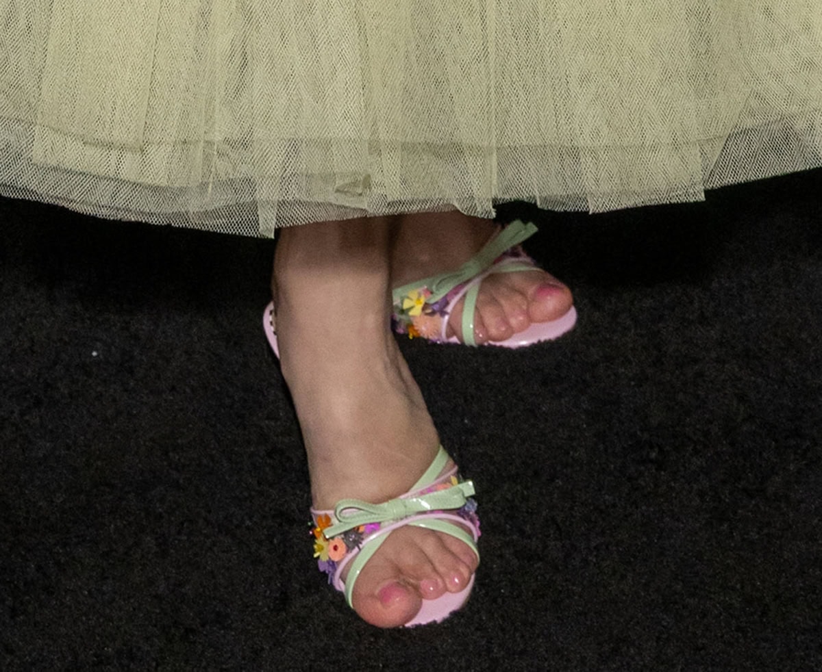 Julia Fox slips her pink-pedicured feet into the Betsey Johnson “Shirley” mules featuring a pink mesh strap with colorful floral appliques, peep toes, a mint green bow, and pink kitten heels