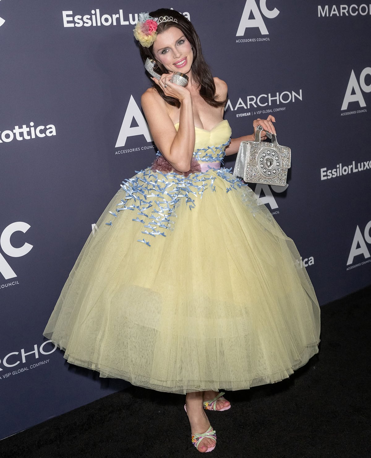 Julia Fox pairs her feminine floral mules with a vintage yellow Betsey Johnson gown featuring a gathered sweetheart neckline, tiny blue ribbons, a pink sash belt with plum floral appliques, and a full, voluminous skirt
