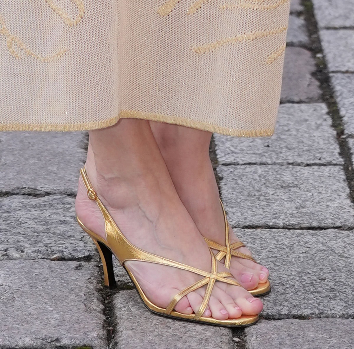 Daisy Lowe wears gold Florrie “Matilda” sandals featuring criss-cross front straps that extend toward the sides and wrap into a slingback strap for a secure, elegant fit