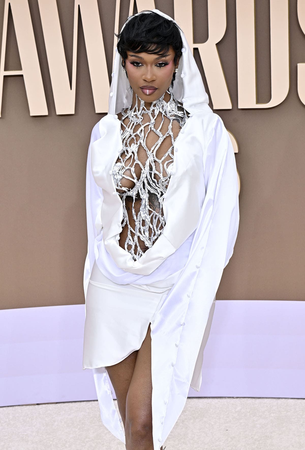 Dess Dior continues with the futuristic cyber-glam theme of her look, pairing her silver gladiators with a silver mini dress by M’kayla Lynne featuring a web-like cage bodice and a white mini dress with a draped front and a hood