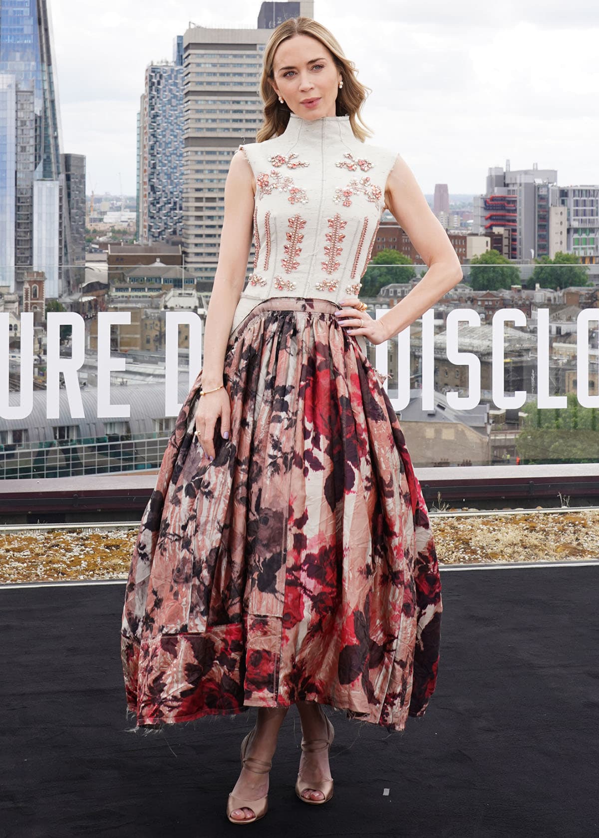 Emily Blunt radiates romantic avant-garde elegance, pairing her soft metallic shoes with an Erdem outfit featuring a structured off-white heathered canvas bodice with pink and red floral crystals and a painterly floral midi skirt with a raw hem