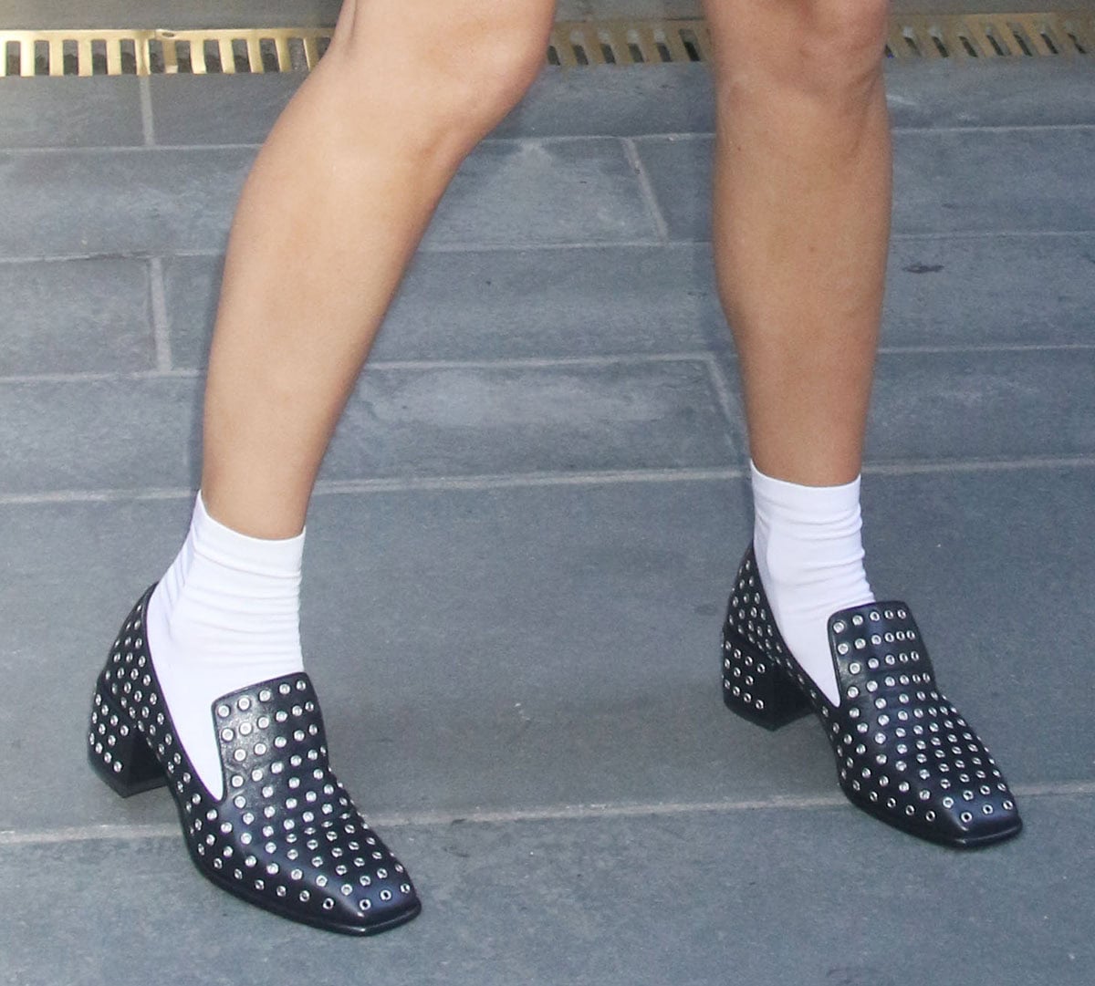 Rashida Jones channels modern schoolgirl energy, wearing white socks with her black Jimmy Choo “Eliot” slipper loafers featuring all-over grommet embellishments, a high-cut vamp, square toes, and short block heels