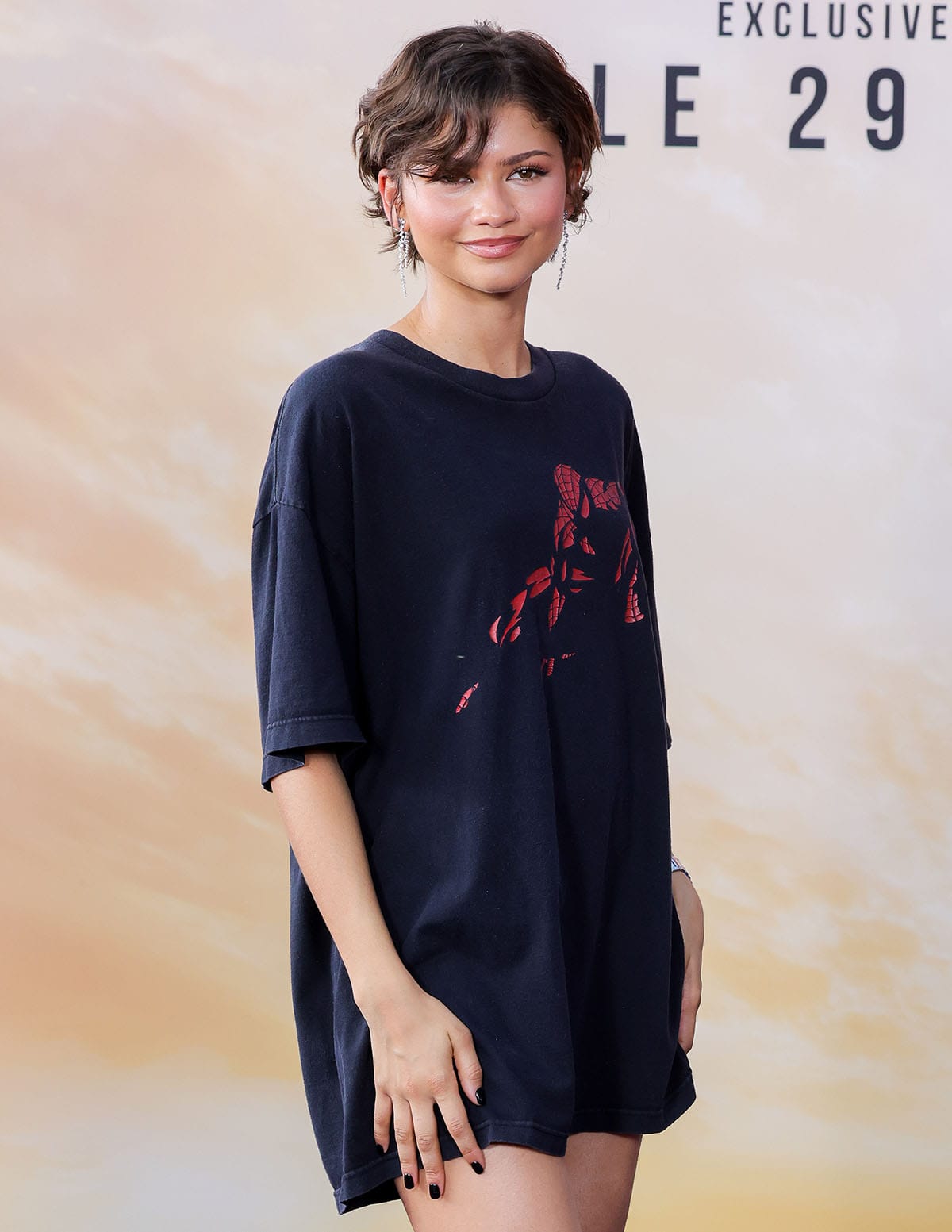Zendaya elevates her laid-back photocall ensemble with dangling earrings, a wavy bixie hairstyle, and pink makeup