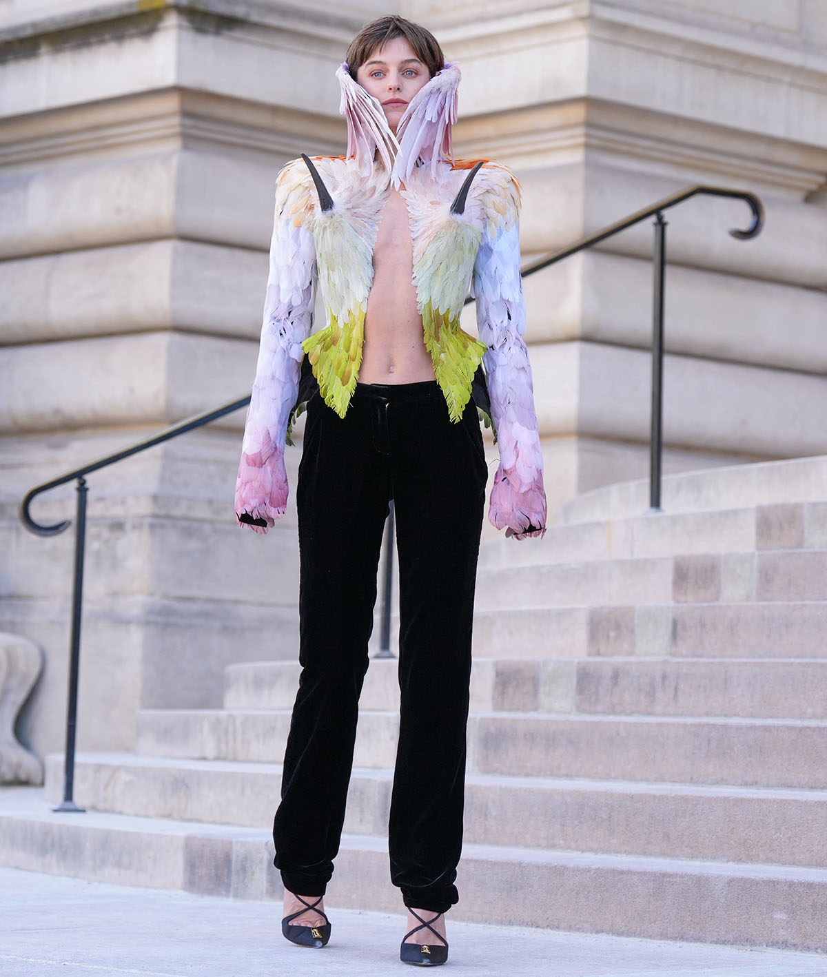 Emma Corrin teams her pumps with black trousers and a jacket covered in pastel ombré plumage, fading from soft purple to icy blue, pale green, yellow, and light orange, black pointed beaks, and a high feather-covered neck