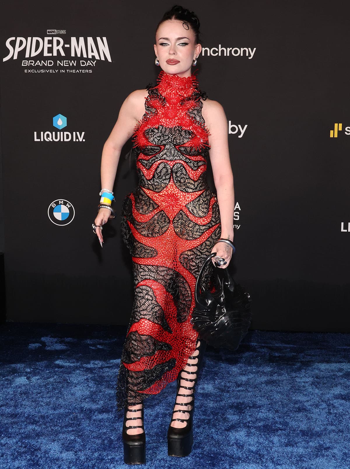 Emma Norton embraces the Spider-Man aesthetic in a black-and-red Karina Bond embroidered lace dress with web-inspired cutouts, paired with towering Marc Jacobs "The Kiki" knee-high platform boots featuring strappy cutouts, chunky platforms, and sky-high block heels
