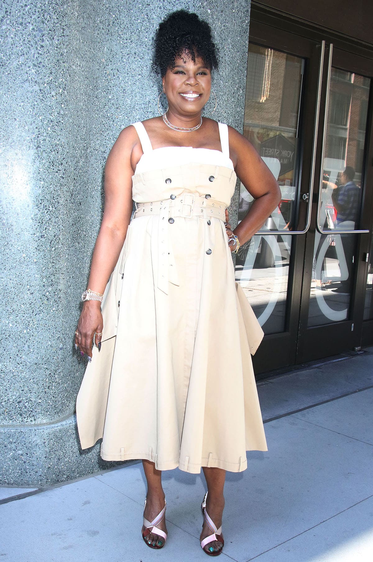 Leslie Jones pairs her multicolored crisscross sandals with a beige Rokh trench dress featuring a double belt, double-breasted button detailing, and belt loops along the neckline and hem