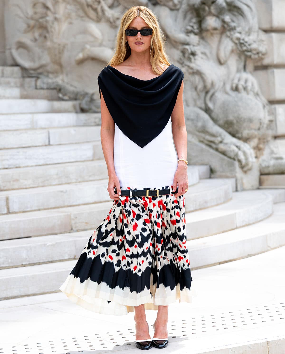 Sarah Pidgeon pairs her glossy two-tone slingback pumps with a Chanel sleeveless dress featuring an integrated black scarf-like bib, a belted drop waist, and a painterly pleated midi skirt
