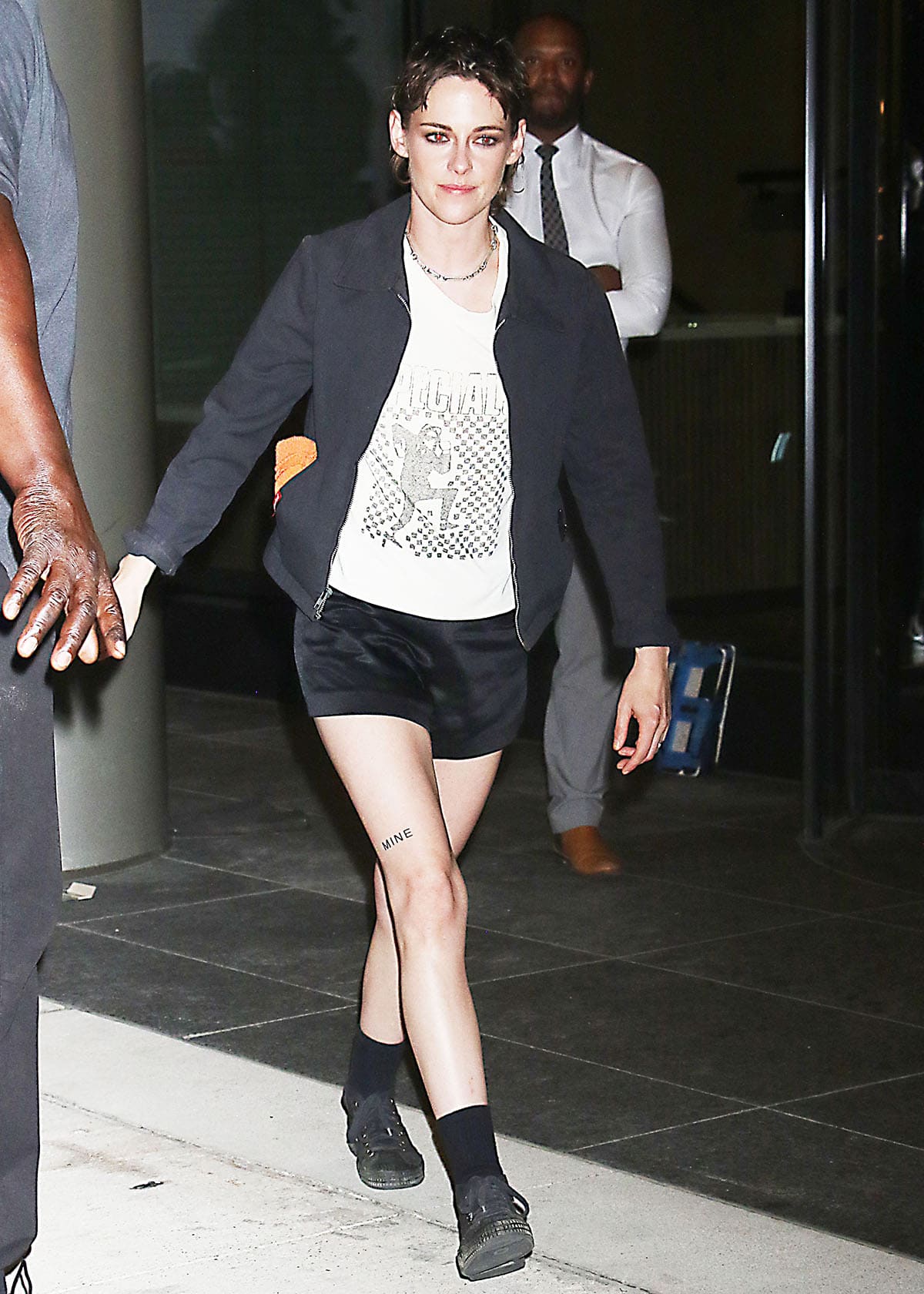 Kristen Stewart embraces her cool, edgy style, pairing her black mid-cut sneakers with black socks, tailored shorts, a black Dickies jacket, and a white The Specials band tee