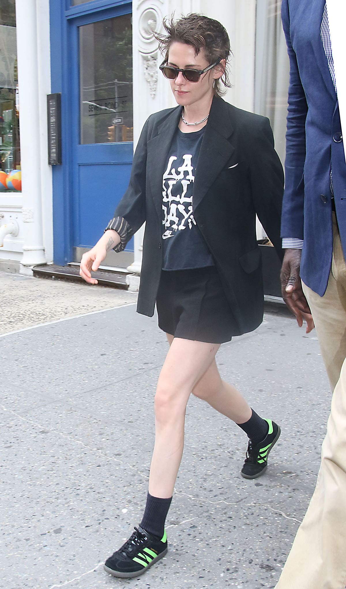 Kristen Stewart gives sharp black tailoring a sporty twist, pairing a boxy blazer and tailored shorts with a cropped Nike “LA All Day” tee and black Adidas Samba Deco SPZL sneakers accented with vivid Lucid Lime stripes