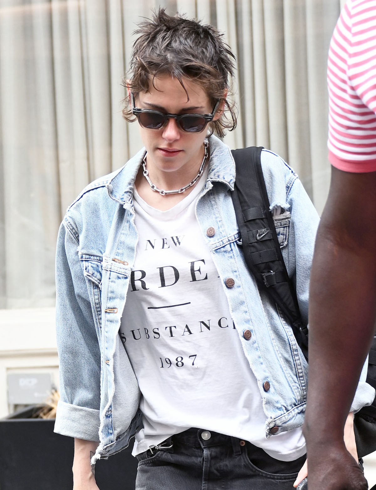 Kristen Stewart layers silver chain jewelry and retro acetate sunglasses over her distressed denim look, with a black Nike backpack adding a practical finish