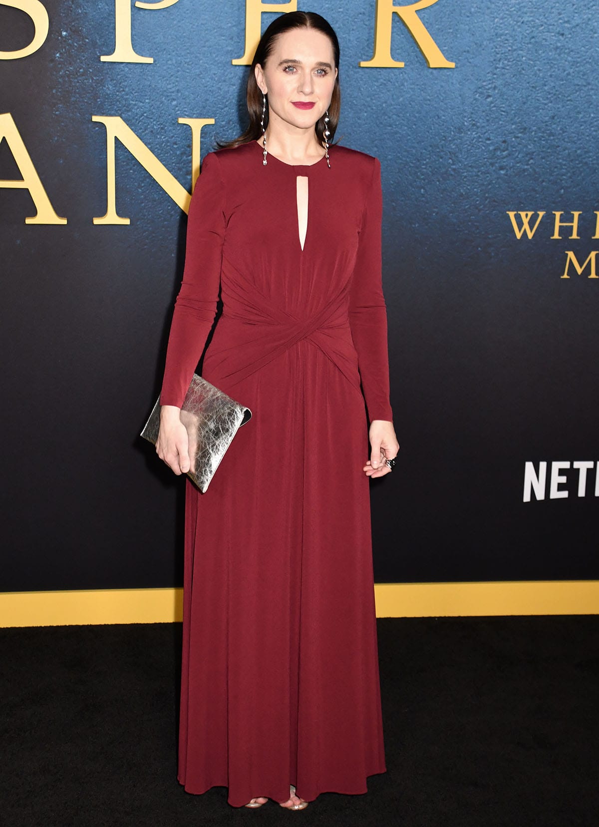 Lena Hall embraces a regal look, pairing her silver heels with a burgundy Nissa dress featuring a keyhole cutout, a twist detail at the waist, long sleeves, and a floor-skimming skirt