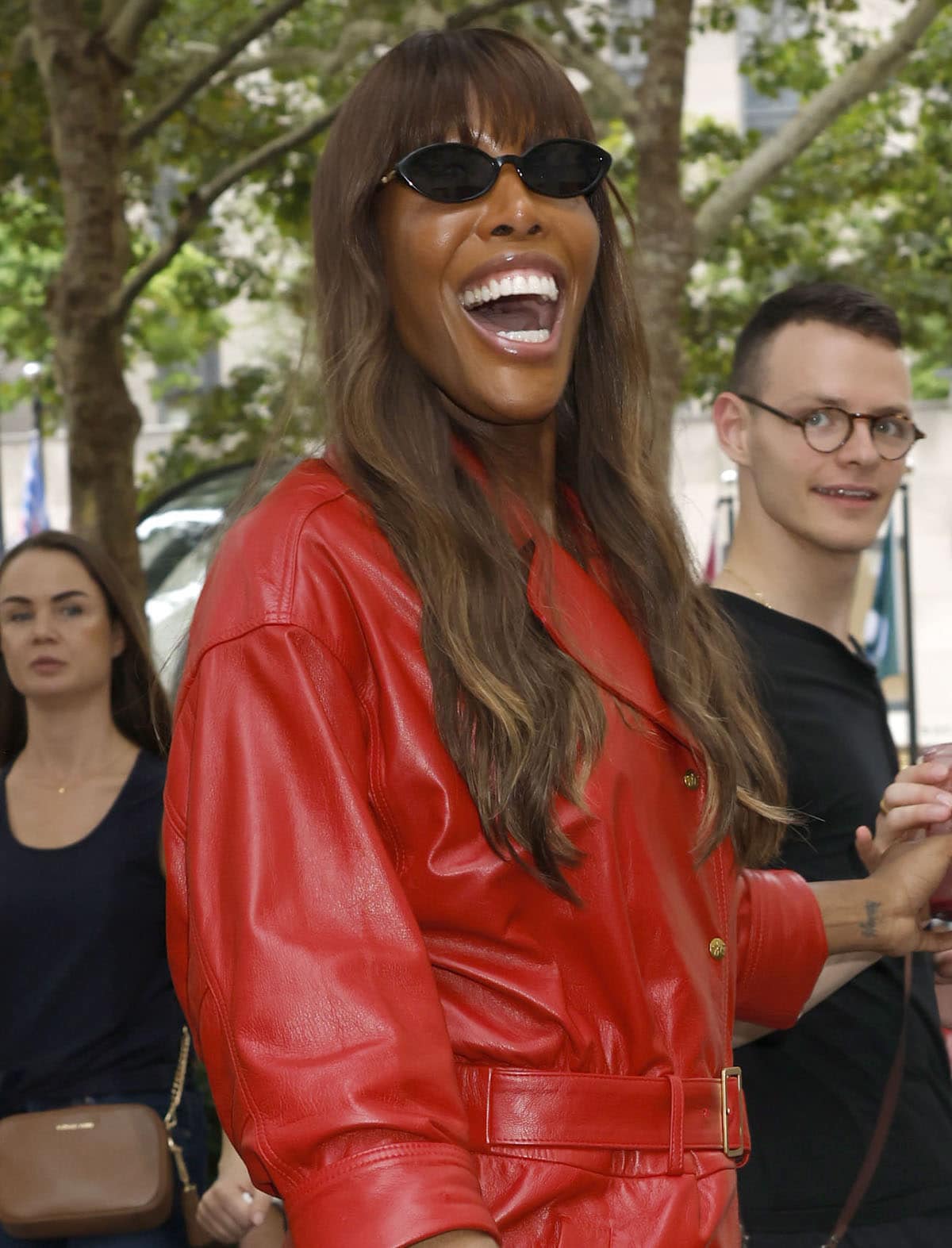 Iman finishes her statement look with sleek black oval sunglasses, glossy makeup, and long brunette waves, letting her cherry-red leather trench coat remain the focus