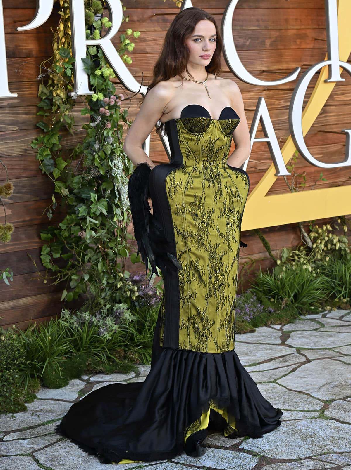 Joey King pairs her pumps with a gothic-romantic gown by Ilya Migmoon featuring chartreuse, slime-green fabric with black lace overlay, exaggerated hip panniers with black wing accents, and a ruffled mermaid hem