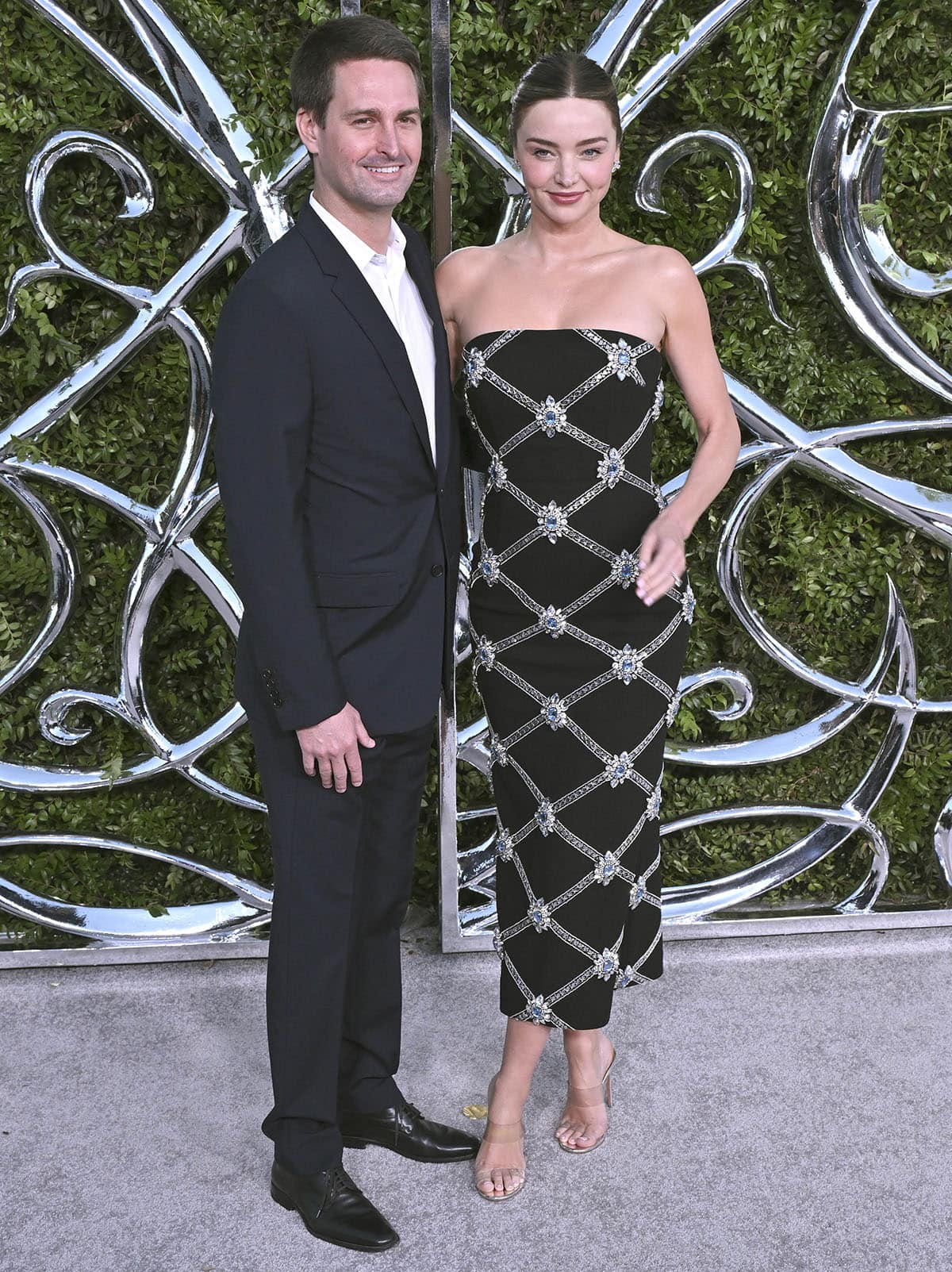 Miranda Kerr wears a black Rebecca Vallance Crown Jewel dress with Aquazzura clear mules alongside Evan Spiegel