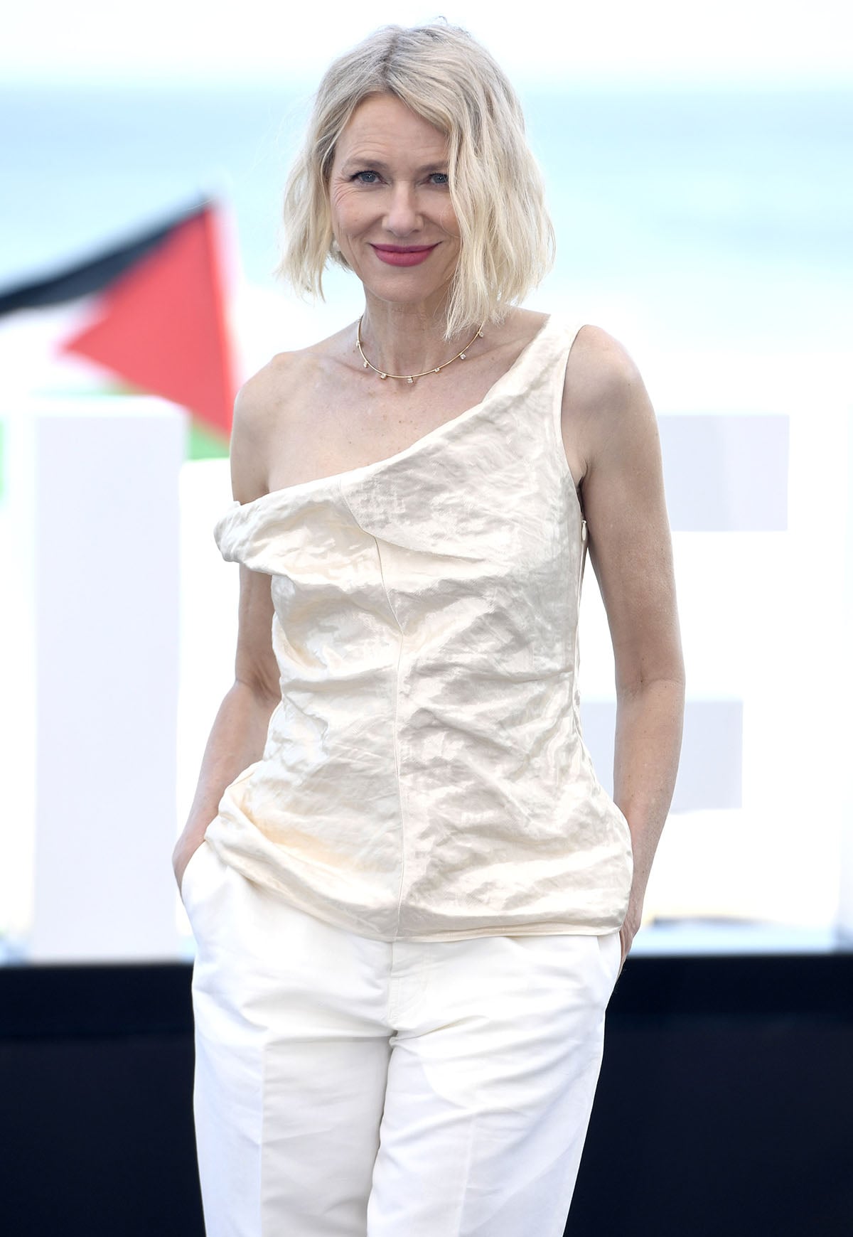 Naomi Watts wears an off-white Fforme one-shoulder top, white wide-leg trousers, and beige python Jude pumps at The Housewife photocall