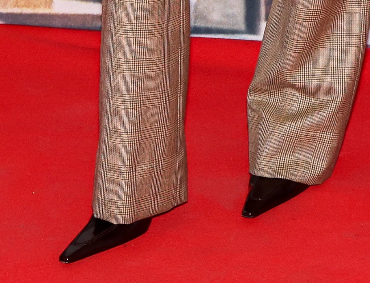 Close-up of Olivia Wilde's glossy black Saint Laurent patent pumps with elongated pointed toes beneath brown checked flared trousers