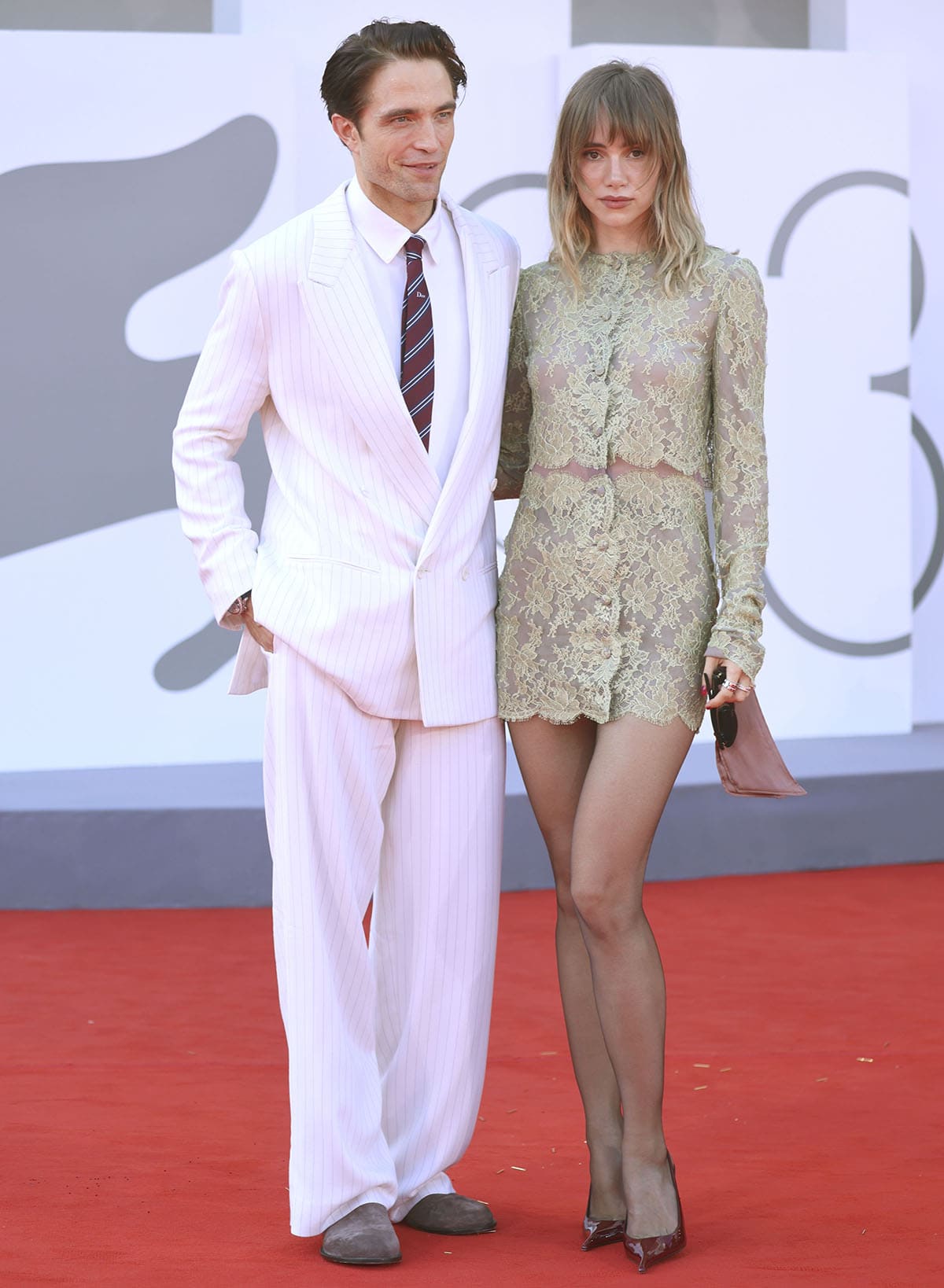 Suki Waterhouse pairs her sage-green Saint Laurent lace mini dress and black tights with burgundy patent Apolline slingback pumps, while Robert Pattinson wears relaxed white pinstripe tailoring