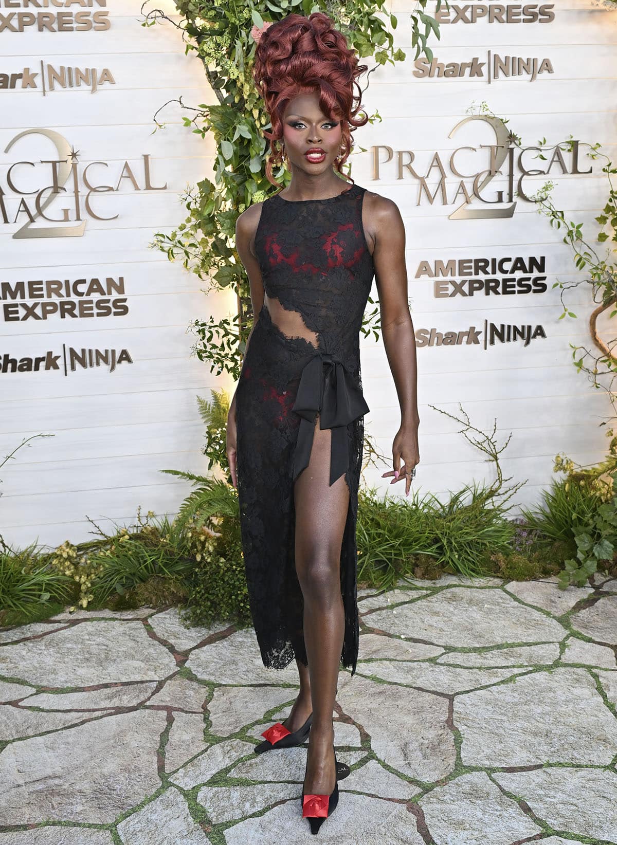 Symone continues the black-and-red theme, pairing her pumps with a sheer black lace dress with a diagonal waist cutout, a side bow, a thigh-high slit, and red underwear
