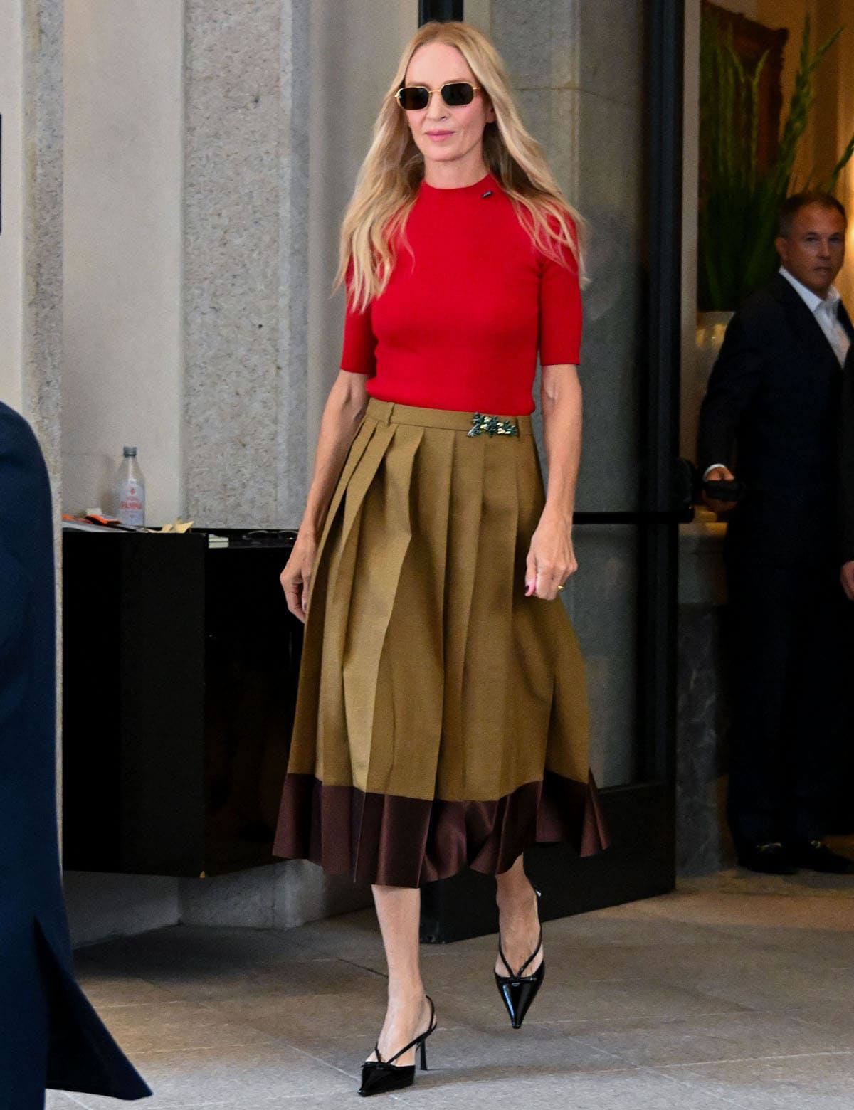 Uma Thurman wears a red Prada short-sleeve sweater with a khaki pleated midi skirt, brown satin hem, crystal brooch, and black slingback pumps
